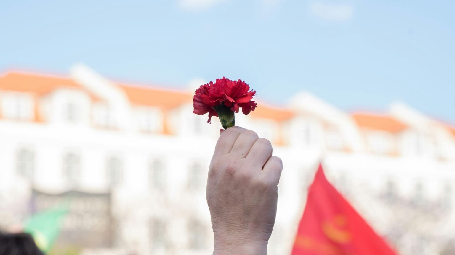 A raised fist holding a single red carnation against a blue sky with blurred white and orange buildings in the background, symbolising Portugal's 1974 Carnation Revolution and Freedom Day on April 25th