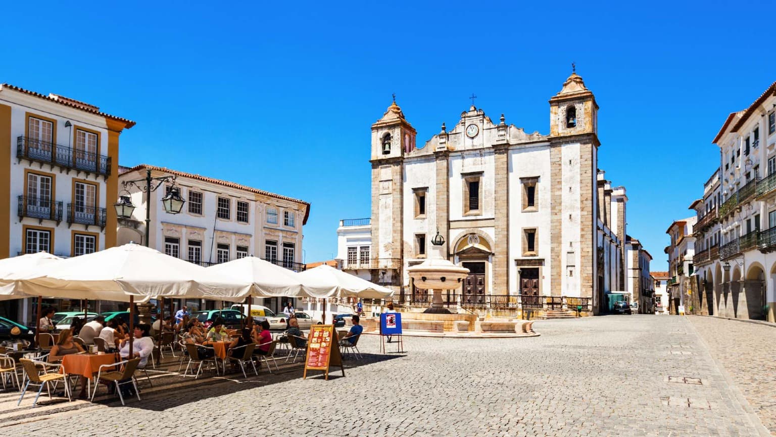 Évora’s historic main square with outdoor cafés and the Church of Santo Antão under a bright blue sky