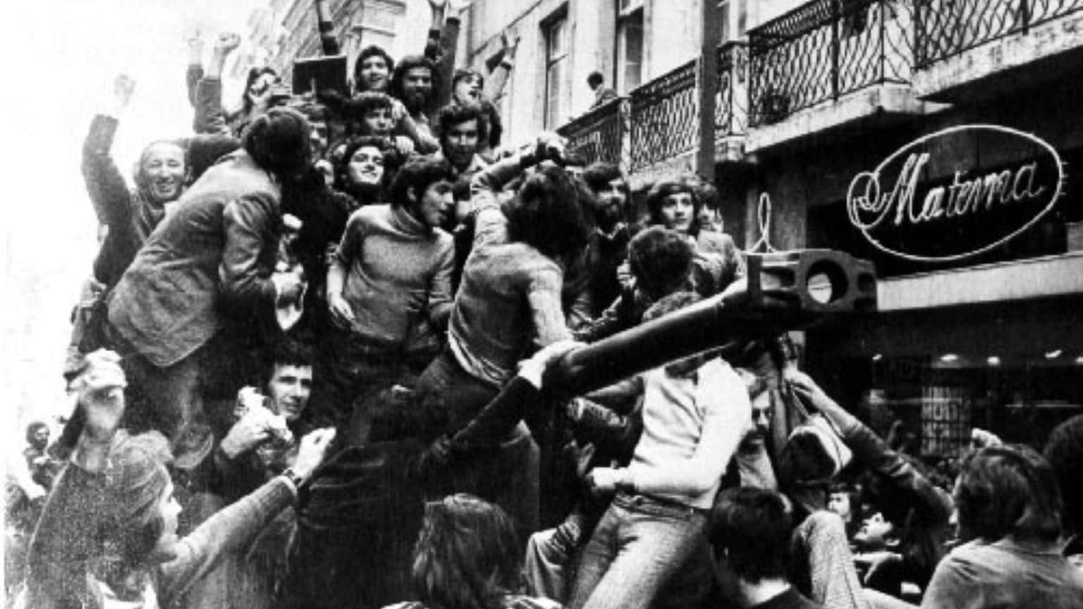 Black and white photograph from April 25 1974 showing jubilant crowds of civilians in Lisbon climbing onto military vehicles and celebrating the Carnation Revolution that ended Portugal's dictatorship