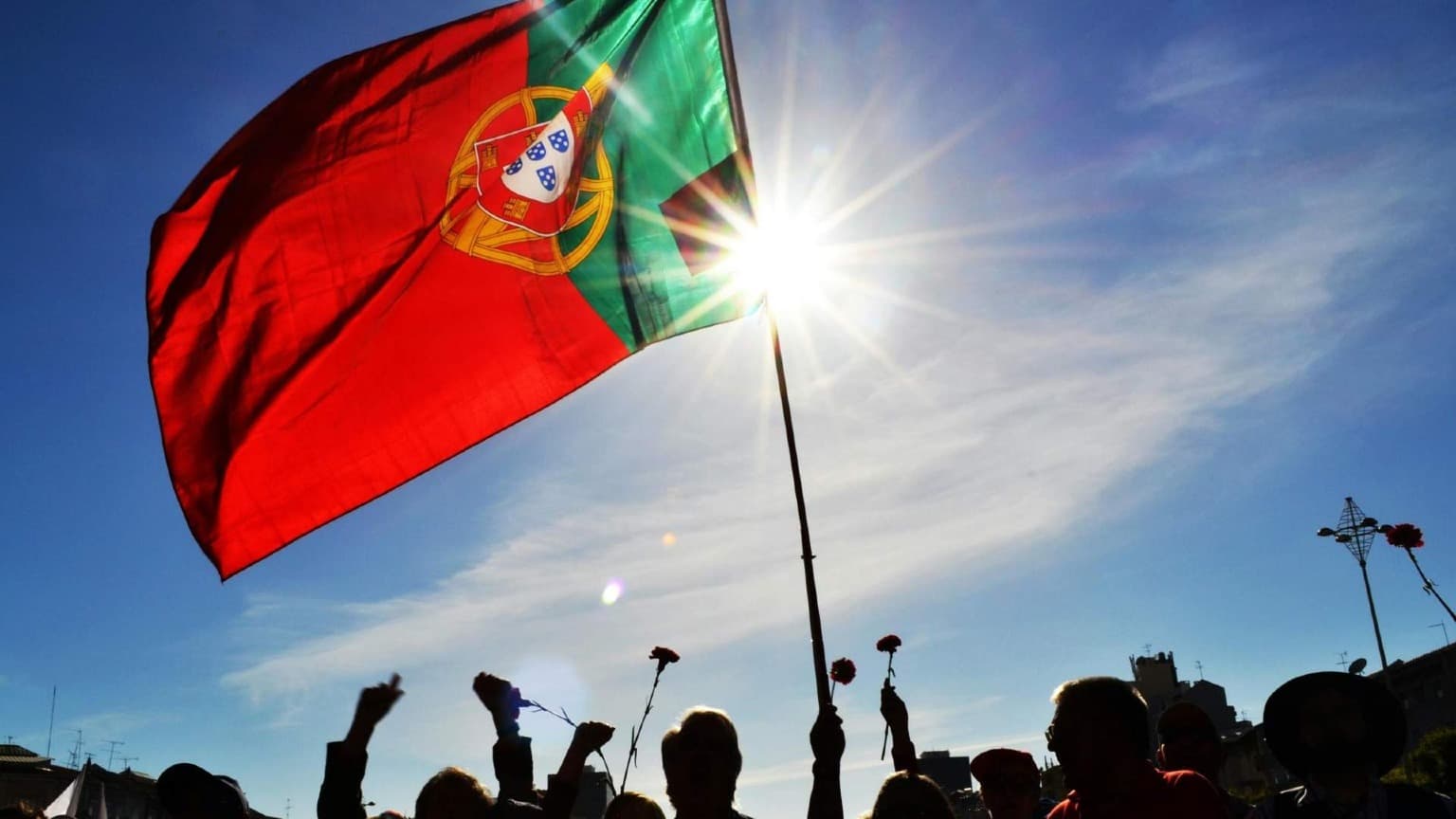 Silhouettes of people holding red carnations and a large Portuguese flag raised on a pole against a bright blue sky at an April 25th Freedom Day celebration in Portugal